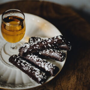 Czekoladowe rurki posypane kokosem na talerzu, obok kieliszek z bursztynowym napojem.