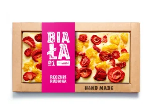 Ręcznie robiona biała czekolada z suszonymi truskawkami i cytrusami w kartonowym opakowaniu z różowym akcentem.