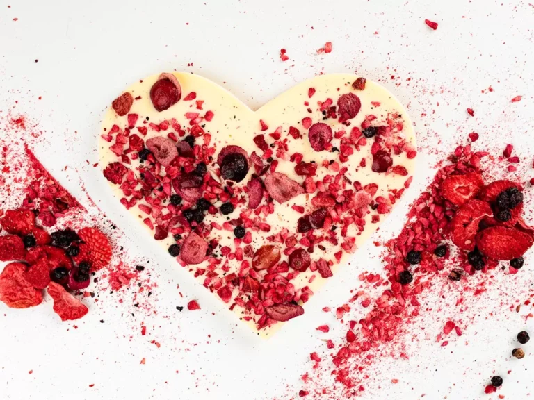 Serce z białej czekolady z kawałkami suszonych owoców na białym tle, otoczone czerwonymi owocami.