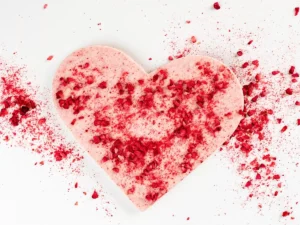 Różowe serce z czekolady posypane suszonymi malinami na białym tle. Romantyczny deser na Walentynki.